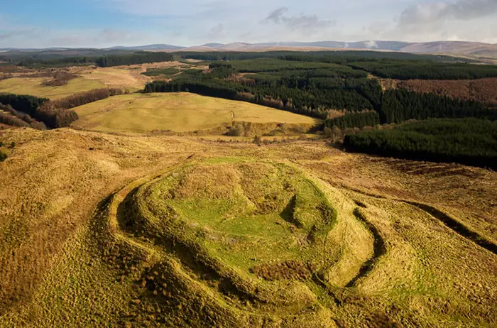 Aerial view of remains of a fort now covered in wild grasses at top of hill with view to land beyond