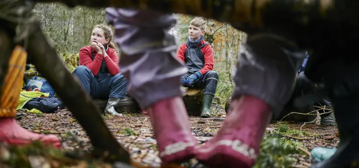 Child's pink wellington boots with purple waterproof trousers as they sit on a log with view of seated children in the background.