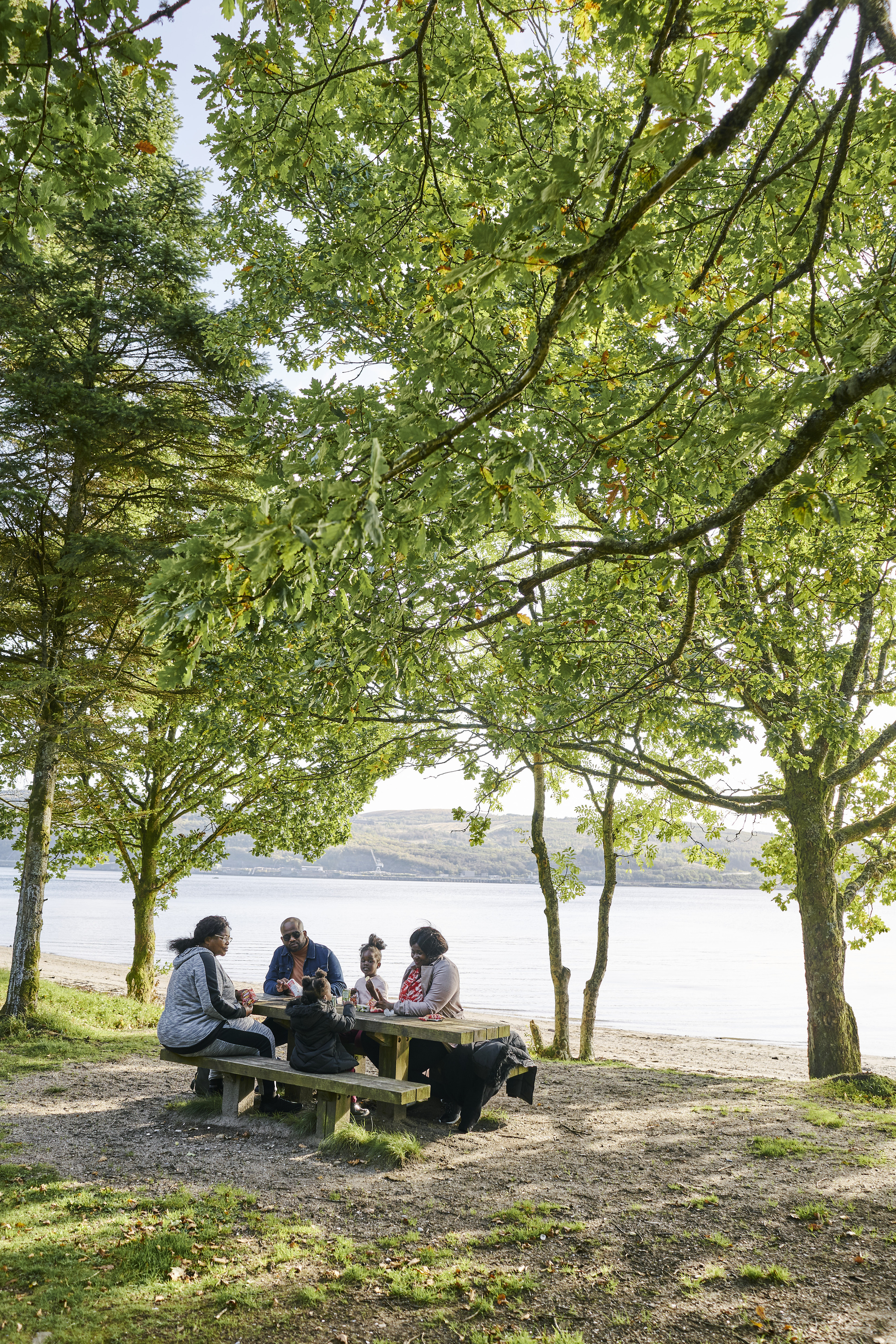 Man, two women and two young girls sit and eat at picnic table, beside beach at Ardentinny, on the shore of Loch Long