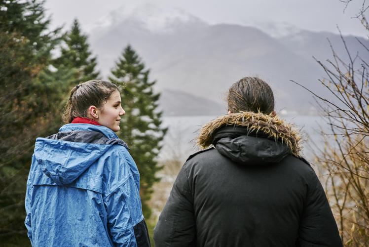 Young woman wearing blue waterproof jacket and young man in black jacket, stand, on a rainy day, at viewpoint near Tom a Chomrain looking over Loch to misty snow capped Pap of Glencoe mountain