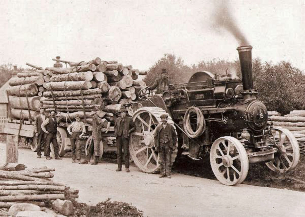 Forestry workers standing in front of traction engine with logs on a trailer on the back.