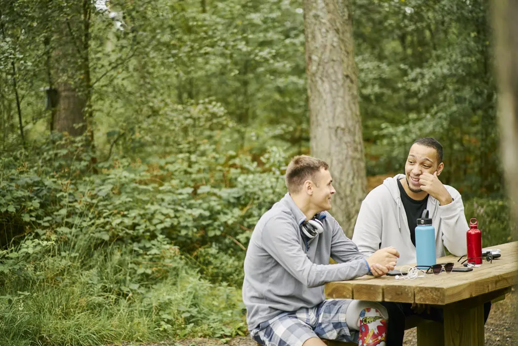 Two men (one with prosthetic leg) sit together chatting at picnic table, on woodland trail, Devilla Forest, near Kincardine.