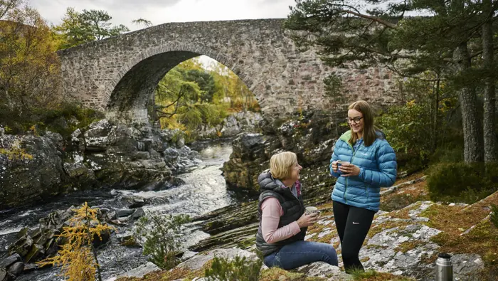 Two people with cups and a flask next to a river that has a bridge over it.