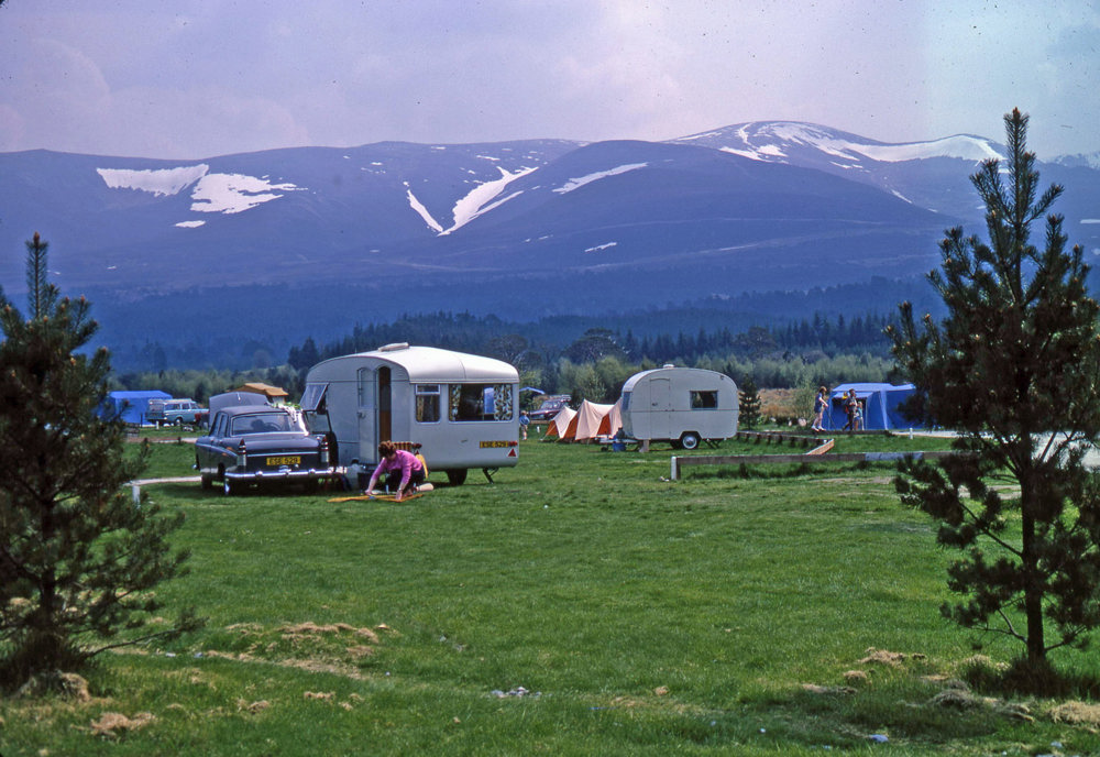 Cars and caravans on a grassy field with mountains in the background.