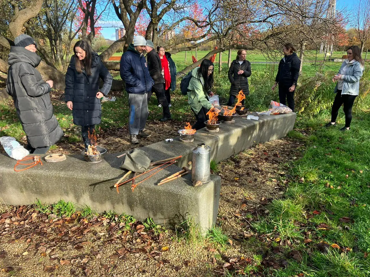 A group of people stand in an urban park setting. Small fires sit on a concrete platform. 