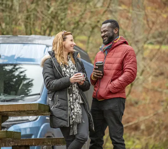 couple standing next to picnic table in front of campervan with reusable travel coffee mugs