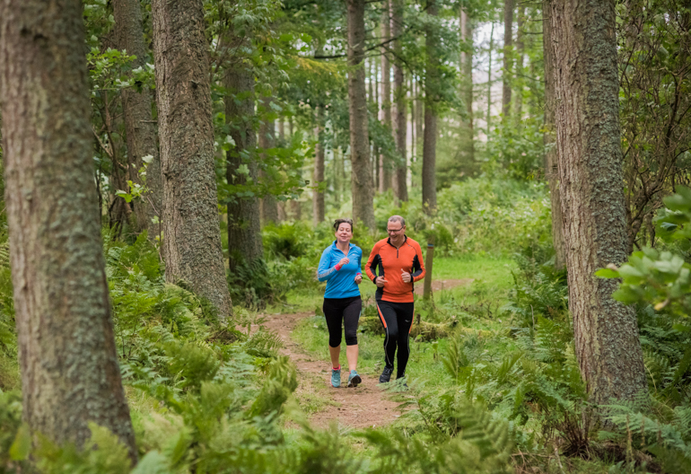Male runner (in orange) and female runner (in blue) run through woodland, Cademuir Forest, near Peebles