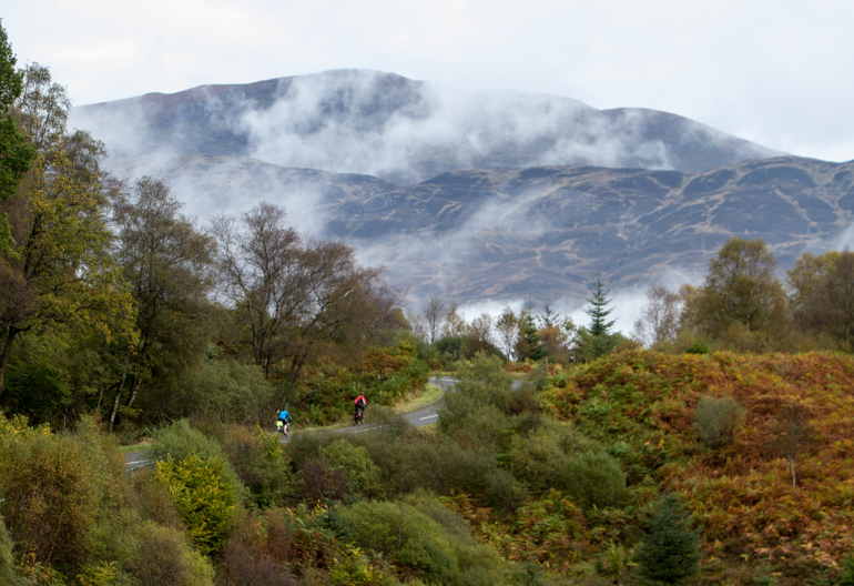 Two cyclists riding gently uphill on a road bordered by green trees and looking out to a large bare hillside with wisps of cloud.