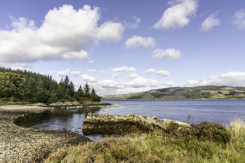 The stone ruins of an old pier in a rocky beach with trees on shore and hills across the water