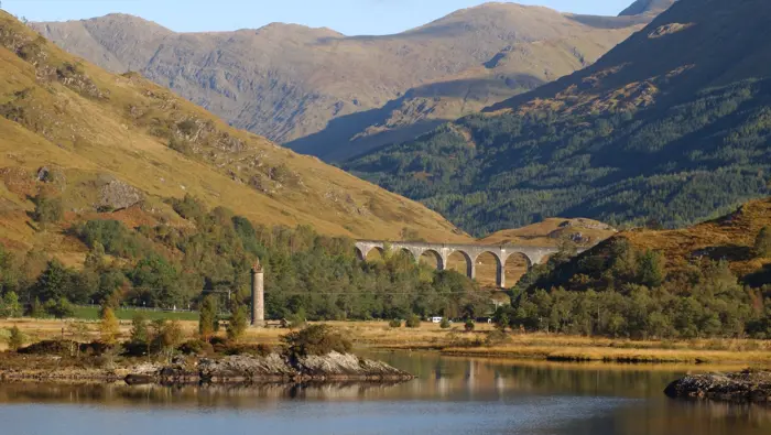 A viaduct and statue along a loch with mountains behind