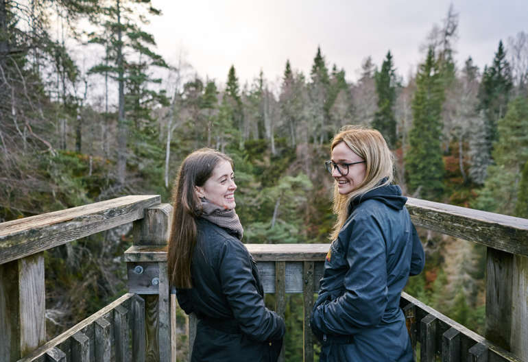Two young women in blue jackets stand, smiling, on the wooden viewing platform overlooking forest of Douglas fir trees and Plodda Falls, Glen Affric