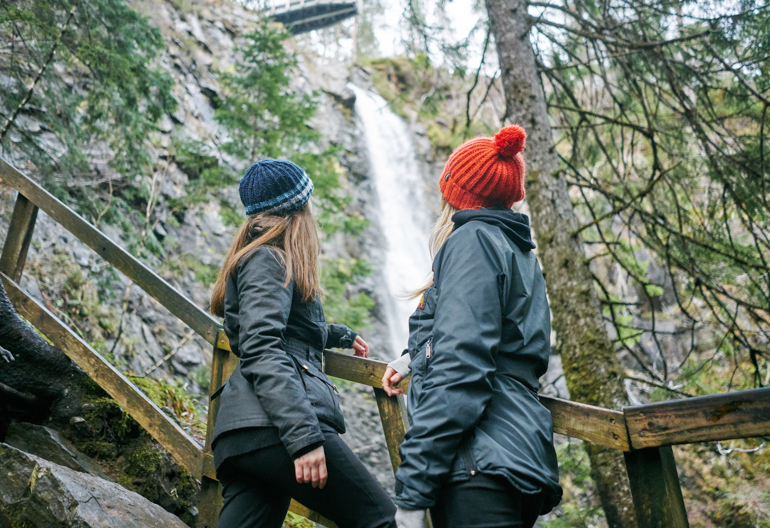 Two young women, one with orange woollen hat, other with blue striped woollen hat, look up from wooden viewing platform to high point of Plodda Falls waterfall as it roars down rockface, Glen Affric
