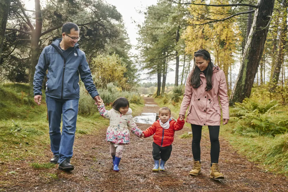 Two adults holding the hands of two small children between them while walking along an off-road forest track among tall trees