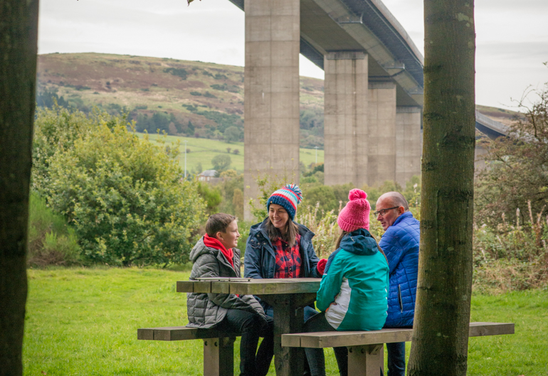 Man, woman, young girl and boy sit together at picnic bench, Boden Boo woodland, Renfrewshire Woods, underneath the Erskine Bridge