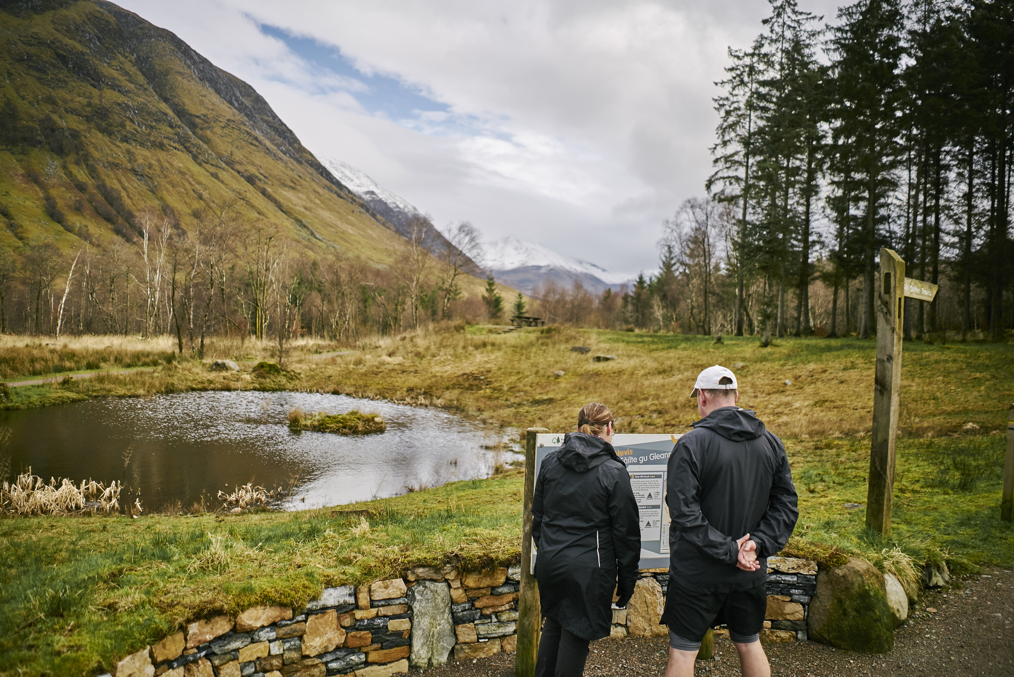 Man and woman in waterproof jackets stand and read interpretation board, with snow capped Ben Nevis in distance, Glen Nevis