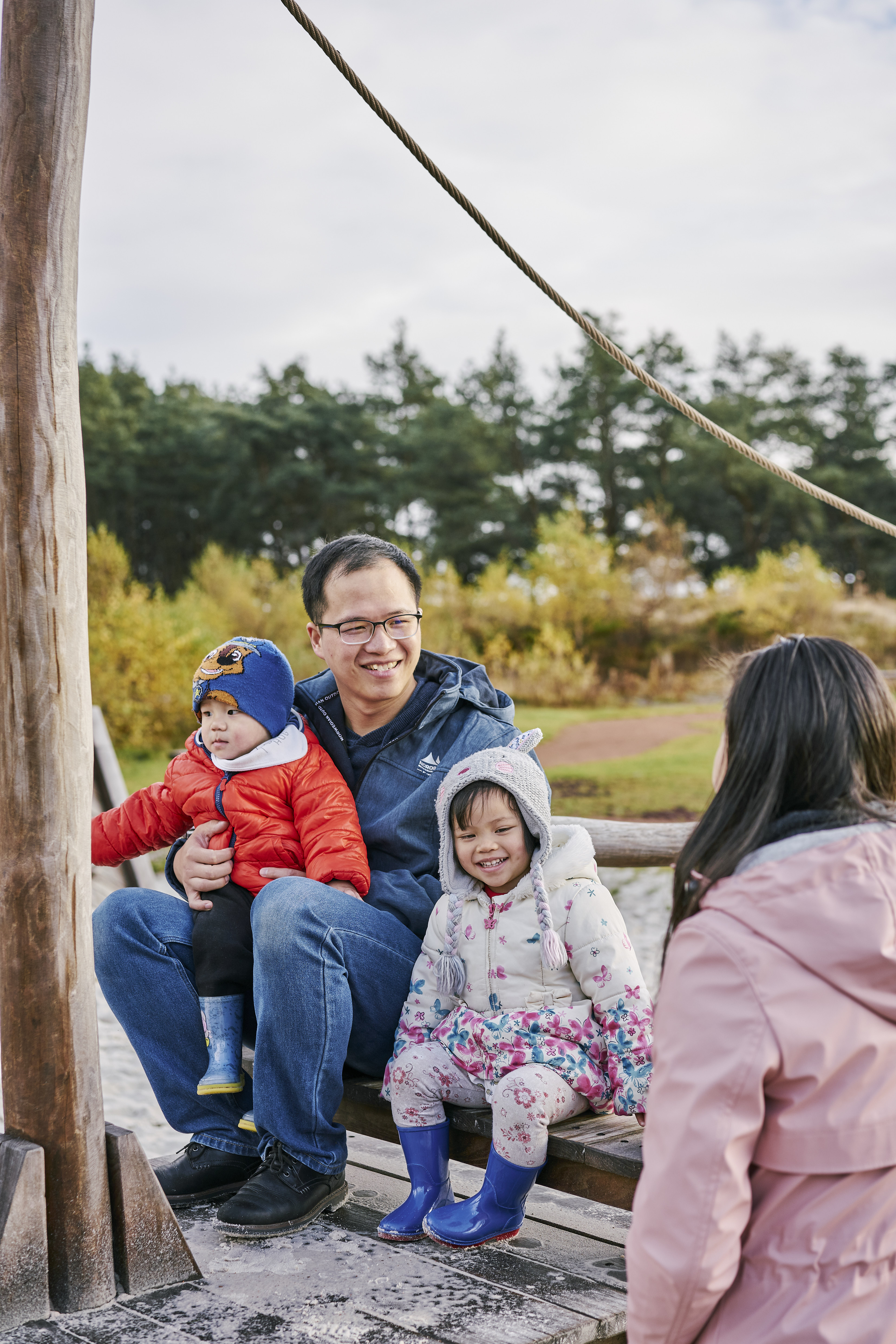 Two young children sitting with man on adventure play construction at Tentsmuir as woman looks on.