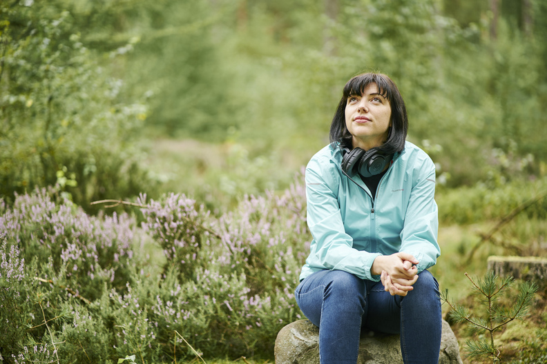 Young woman in blue jacket sits in woodland, looking into distance, Devilla Forest, near Kincardine