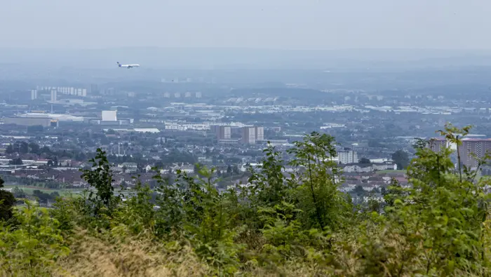 View of the city with green shrubs and trees in the foreground and a plane flying overhead.