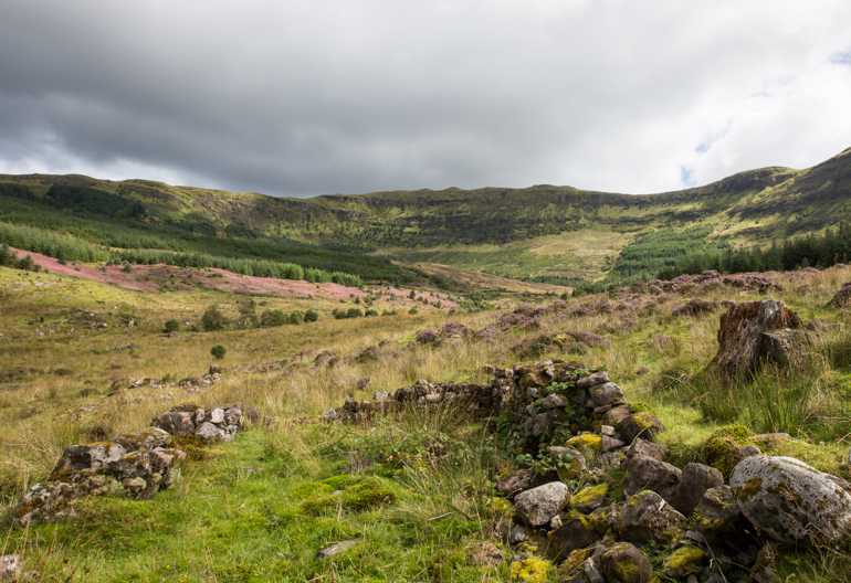 Heather covered ground with rocks and grass covering the ruins of a Highland village with a mountain rage behind