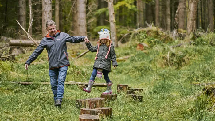  Man helps young girl walk on wooden stepping stones, on the balance trail, Upper Reelig Community Wood, near Inverness.