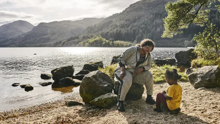 Woman and young girl playing together with bucket and sand, on shoreline at Loch Eck, with loch and surrounding hills in background