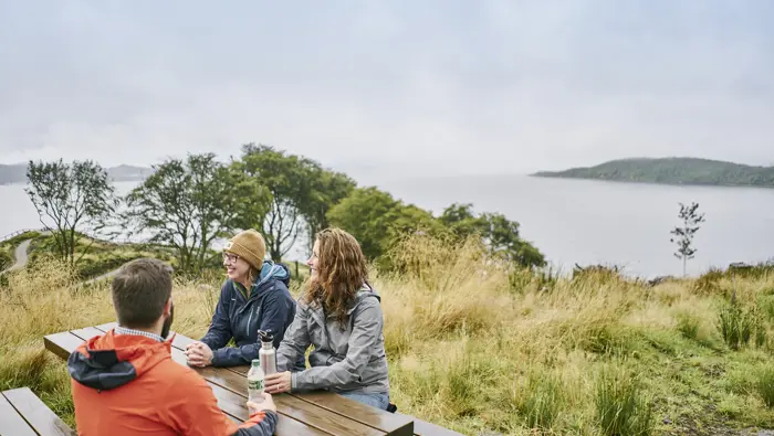 Three people sitting around a picnic bench with views out to sea in the background.