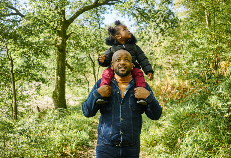 Man with young girl on his shoulders walks through leafy track, beside beach at Ardentinny, on the shore of Loch Long