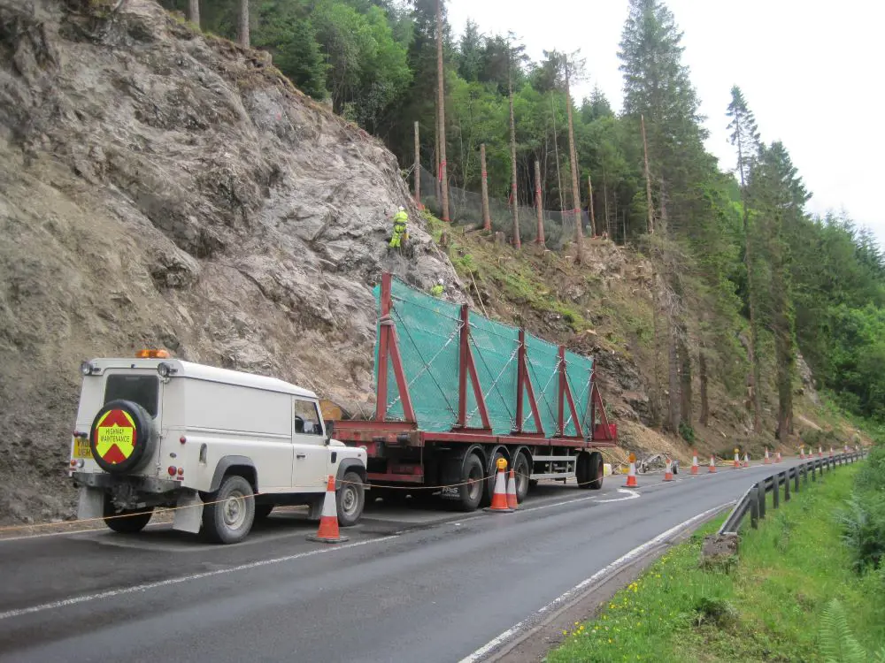 A white jeep and truck bed next to a steep hillside with workers in safety gear propelling from the rock